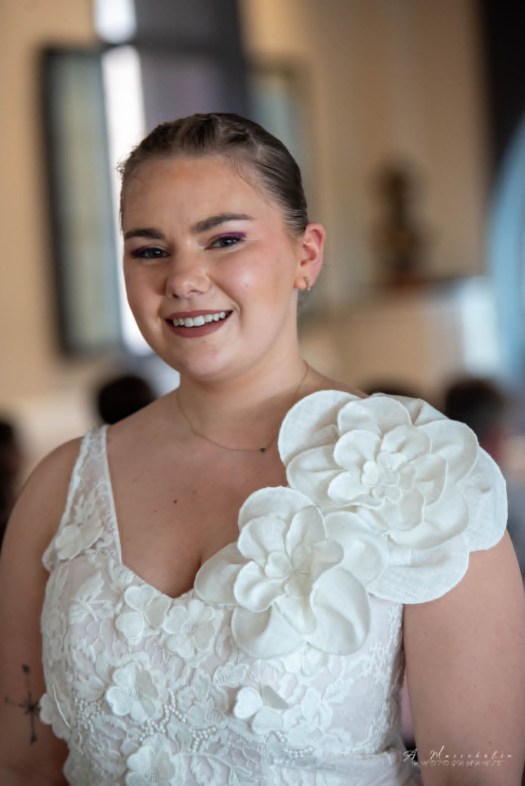Femme souriante portant une robe blanche ornée de grandes fleurs en tissu, dans un cadre intérieur lumineux.