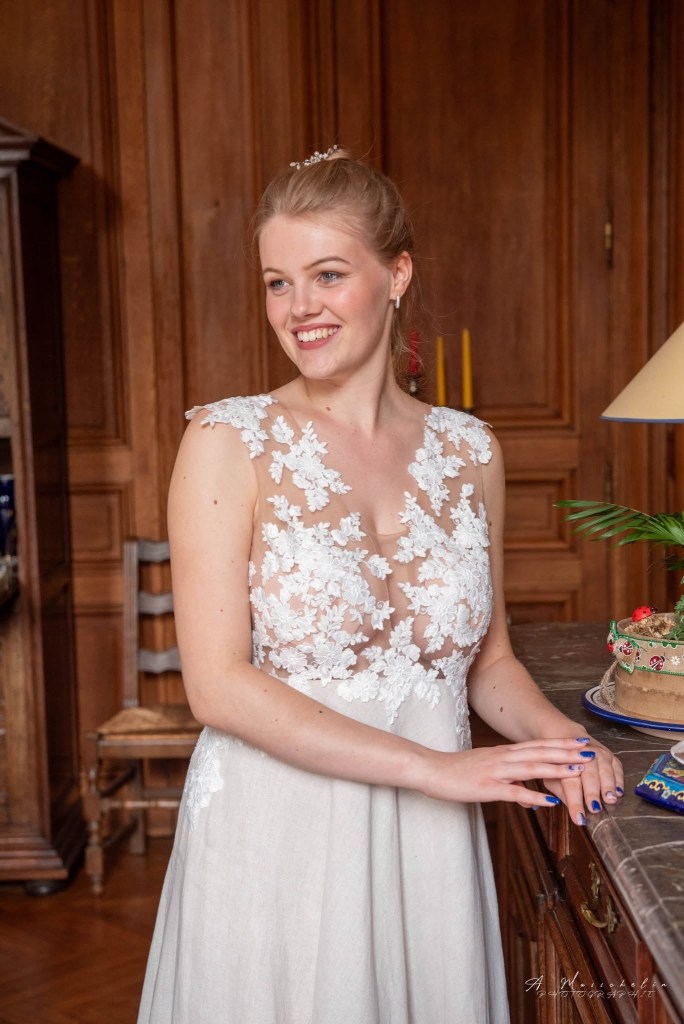 Femme souriante en robe de mariée avec des appliqués floraux, posant devant une table en bois dans une pièce bien éclairée.