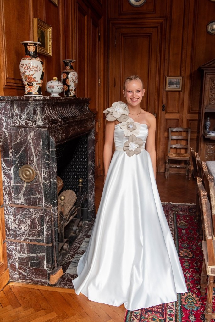 Une jeune femme souriante porte une robe blanche élégante avec des fleurs décoratives, se tenant devant une cheminée en marbre dans un intérieur en bois, orné de vases et de meubles anciens.